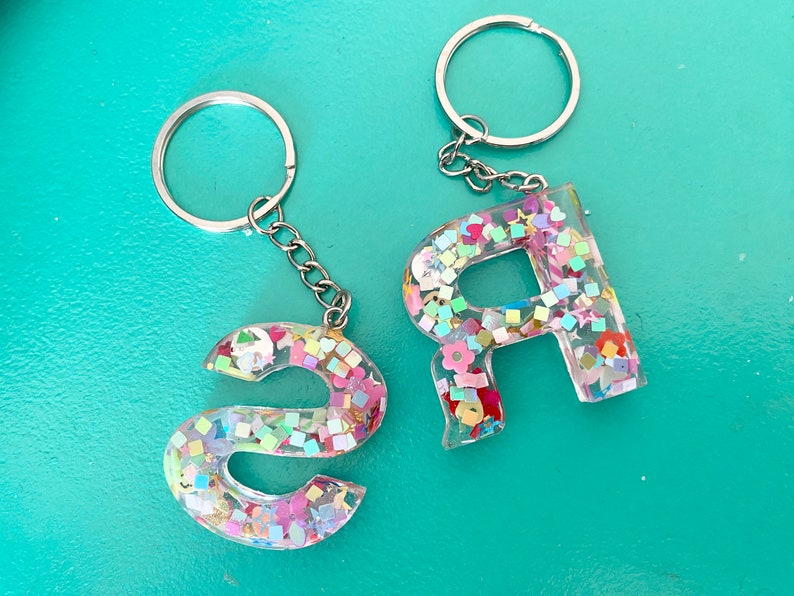 May include: Two keychains with silver key rings and chains. The charms are clear resin letters filled with colorful glitter and confetti. The letters are "S" and "R".