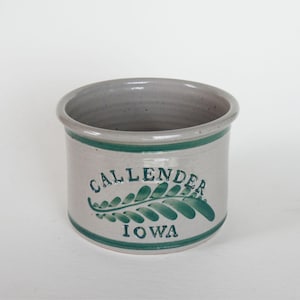May include: A cylindrical ceramic crock with a light gray speckled finish. The exterior features dark green horizontal bands at the top and bottom, with the text CALLENDER and IOWA flanking a central green fern leaf illustration.