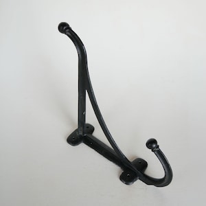 May include: A black metal wall hook featuring a tall vertical arm and a shorter lower hook, both ending in rounded ball finials. The base plate includes four pre-drilled screw holes for mounting, and the structure is connected by a curved support.