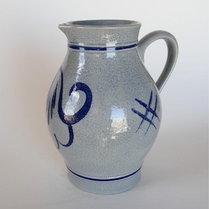 May include: A gray ceramic pitcher with a handle and spout. The pitcher has blue painted designs and accents. The body of the pitcher is textured. The pitcher is likely used for serving beverages.