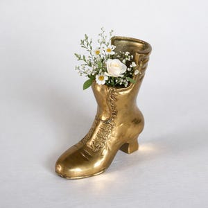 May include: A gold-colored boot-shaped vase filled with white roses, daisies, and greenery. The vase has a vintage design with laces and a small heel. The flowers add a touch of freshness to the decorative piece.