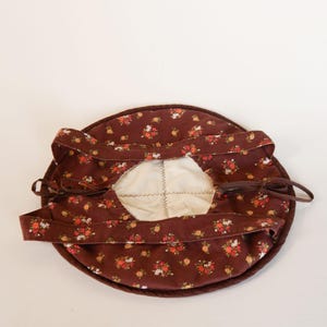 May include: A brown floral print bonnet with a white interior and brown ties. The bonnet features a repeating pattern of small flowers in shades of red, orange, and yellow. The ties are a solid brown color.