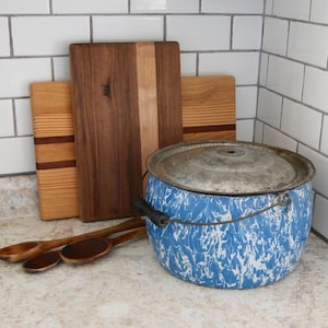 May include: A blue and white enamelware pot with a metal lid, black handle, and wire handle. Three wooden cutting boards and three wooden spoons are behind the pot. The cutting boards are various sizes and wood tones.