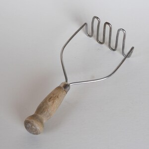 May include: A vintage potato masher with a wooden handle and a stainless steel mashing head. The handle is light brown, and the mashing head has a wavy design. The kitchen utensil is designed for mashing cooked potatoes.
