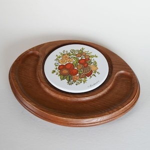 May include: A wooden serving tray with a central circular inset featuring a white ceramic tile. The tile is decorated with a colorful floral and fruit design. The tray has two curved side compartments.