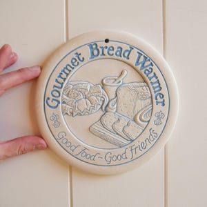 May include: A round, off-white ceramic bread warmer with blue lettering and illustrations. The text reads "Gourmet Bread Warmer" and "Good Food - Good Friends." The center features images of bread, a mortar and pestle, and a cutting board.