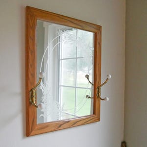 May include: A rectangular wall mirror with a medium-toned wood frame. The glass features an etched floral wreath design. Two brass-toned metal coat hooks with white ball-tipped ends are mounted on the left and right sides of the wooden frame.