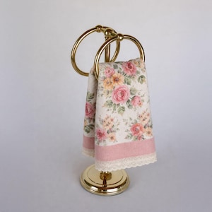 May include: A gold-toned towel holder with two rings, holding a floral-patterned towel. The towel features pink roses and other flowers on a cream background, with a pink border and lace trim. The holder has a round base.