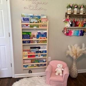 May include: A wooden bookshelf with multiple shelves filled with children's books. The bookshelf is mounted on a light gray wall. The wall also has a pink neon sign that reads "once upon a time..."  A pink armchair with a teddy bear sitting on it is in the foreground.