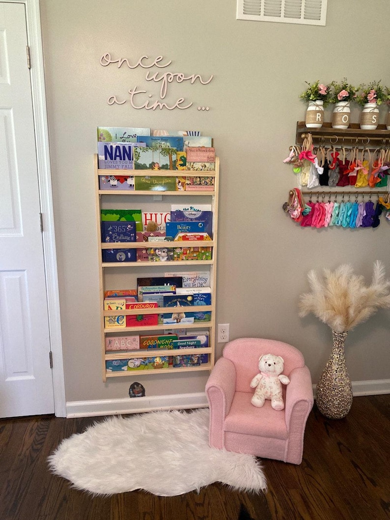 May include: A wooden bookshelf with multiple shelves filled with children's books. The bookshelf is mounted on a light gray wall. The wall also has a pink neon sign that reads "once upon a time..."  A pink armchair with a teddy bear sitting on it is in the foreground.