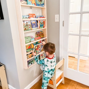 May include: A young child in green and white pajamas reaches for a book on a wooden bookshelf. The bookshelf is mounted on a wall and has five shelves filled with colorful children's books. The child is standing on a wooden step stool.