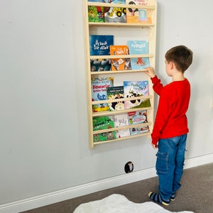 May include: A wooden bookshelf with multiple shelves, filled with colorful children's books. A young boy is standing in front of the bookshelf, reaching for a book. The bookshelf is mounted on a white wall.