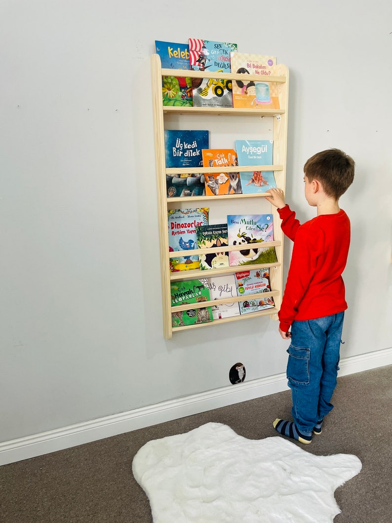 May include: A wooden bookshelf with multiple shelves, filled with colorful children's books. A young boy is standing in front of the bookshelf, reaching for a book. The bookshelf is mounted on a white wall.