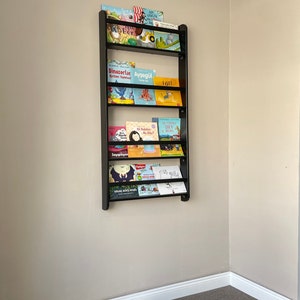 May include: A black metal bookshelf with multiple shelves holding a variety of children's books. The books have colorful covers with illustrations and text in Turkish. The bookshelf is mounted on a light beige wall.