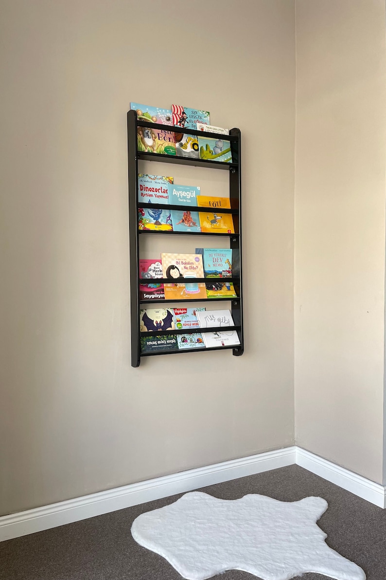 May include: A black metal bookshelf with multiple shelves holding a variety of children's books. The books have colorful covers with illustrations and text in Turkish. The bookshelf is mounted on a light beige wall.