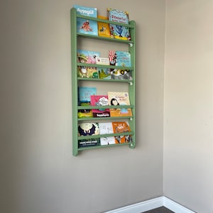 May include: A green wooden bookshelf with multiple shelves filled with colorful children's books. The books have titles in Turkish, including "Aysegul", "Dinozorlar", "Benim Yuregim", "Saygilayim", "Mis Gidi", and "Go Talk! Hayvanlar".