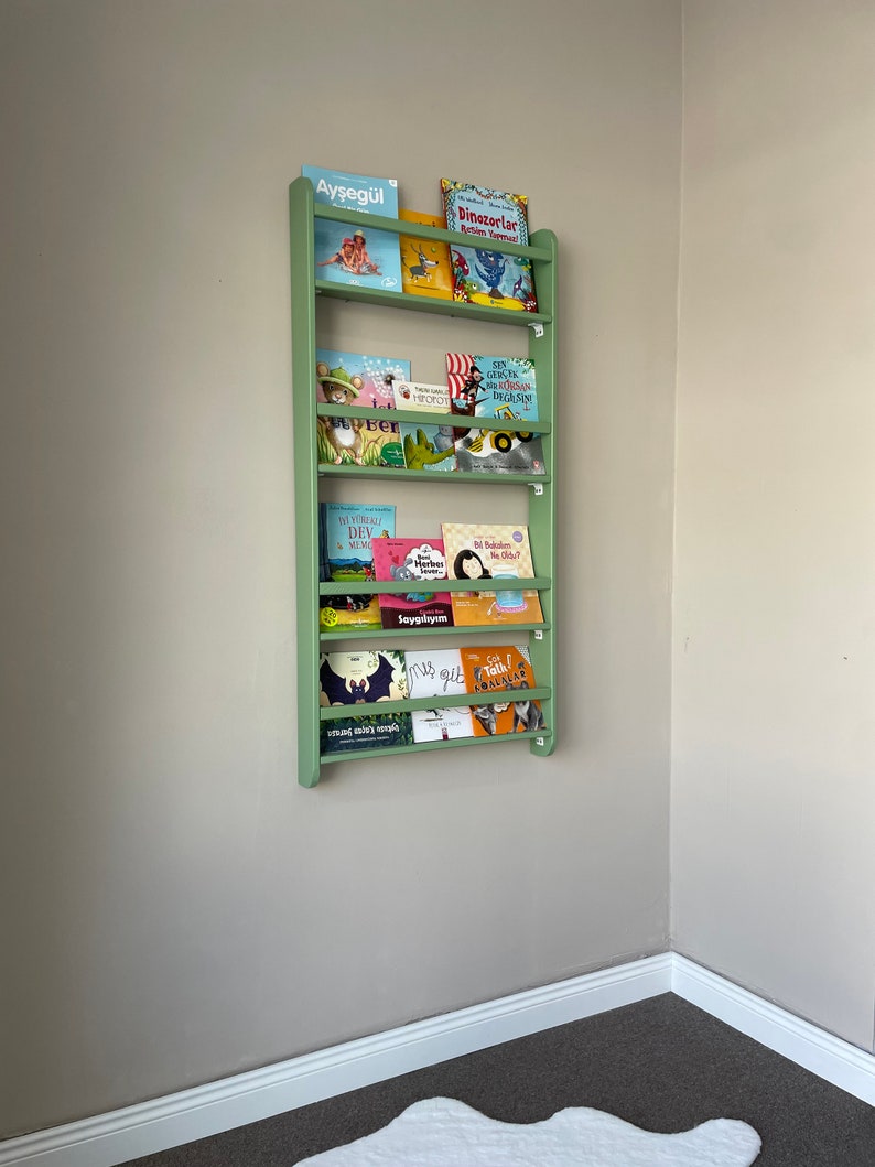 May include: A green wooden bookshelf with multiple shelves filled with colorful children's books. The books have titles in Turkish, including "Aysegul", "Dinozorlar", "Benim Yuregim", "Saygilayim", "Mis Gidi", and "Go Talk! Hayvanlar".