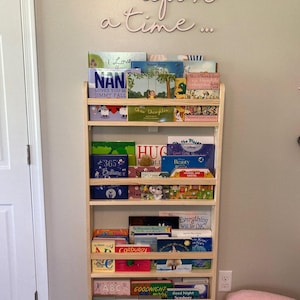 May include: A wooden bookshelf with five shelves filled with children's books. The bookshelf is mounted on a wall painted a light beige color. The words "once upon a time..." are written in pink cursive letters on the wall above the bookshelf.