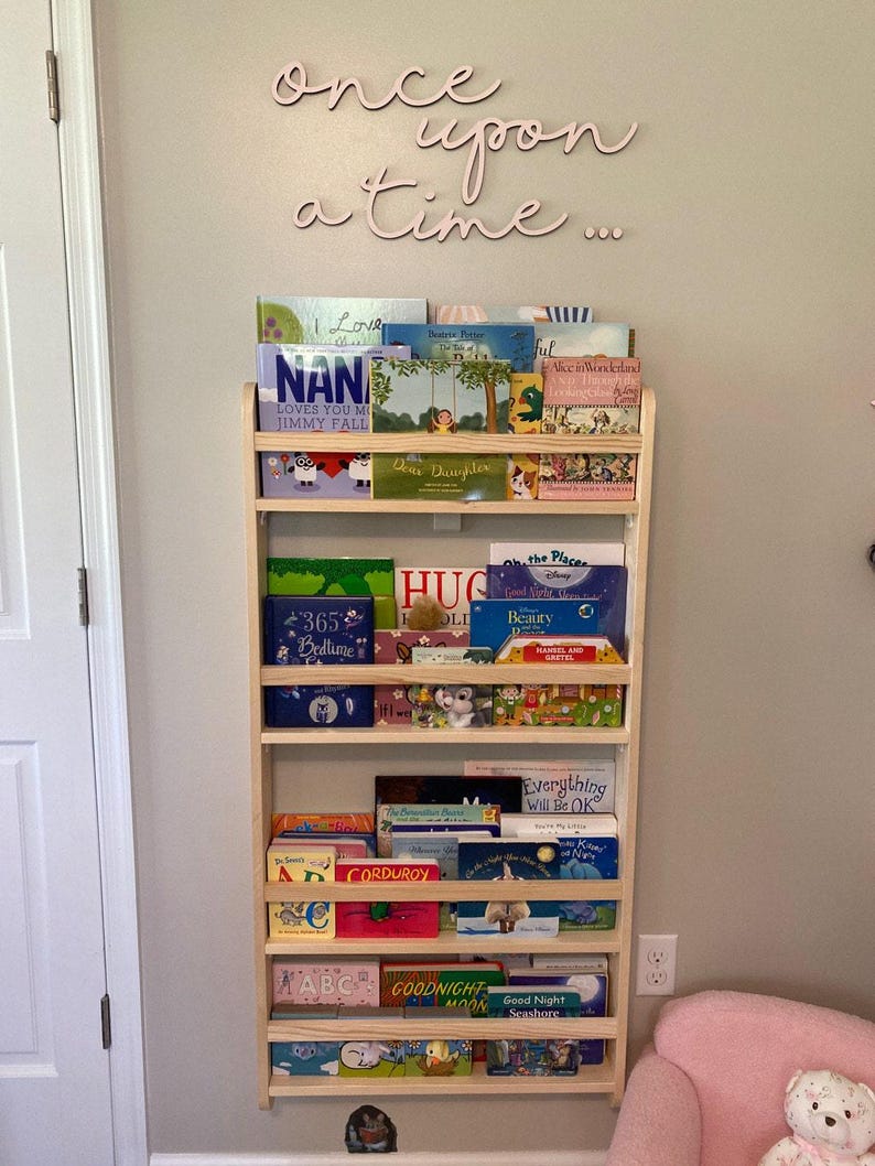 May include: A wooden bookshelf with five shelves filled with children's books. The bookshelf is mounted on a wall painted a light beige color. The words "once upon a time..." are written in pink cursive letters on the wall above the bookshelf.