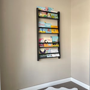 May include: A black metal bookshelf with multiple shelves holds a variety of children's books with colorful covers. The bookshelf is mounted on a light beige wall. A white fluffy rug is on the floor below the bookshelf.