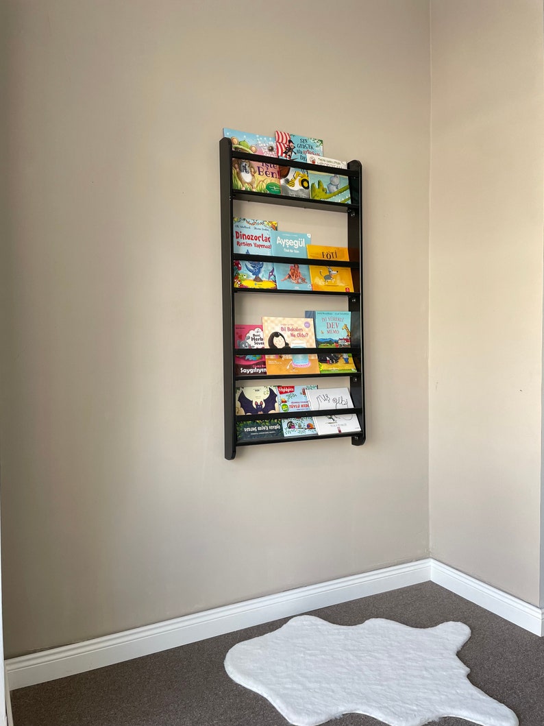 May include: A black metal bookshelf with multiple shelves holds a variety of children's books with colorful covers. The bookshelf is mounted on a light beige wall. A white fluffy rug is on the floor below the bookshelf.