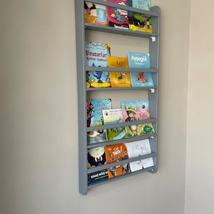 May include: A gray wooden bookshelf with multiple shelves, each holding a variety of colorful children's books. The books have titles in Turkish, including "Bil Bakalım", "Dinozorlar", "P&Ouml;TI", "Ayşeg&uuml;l", "SEN GER&Ccedil;EK BİR KORSAN DEG", "İşte", "&Ccedil;ok Tatlı KonalaLAR", and "us gibi".