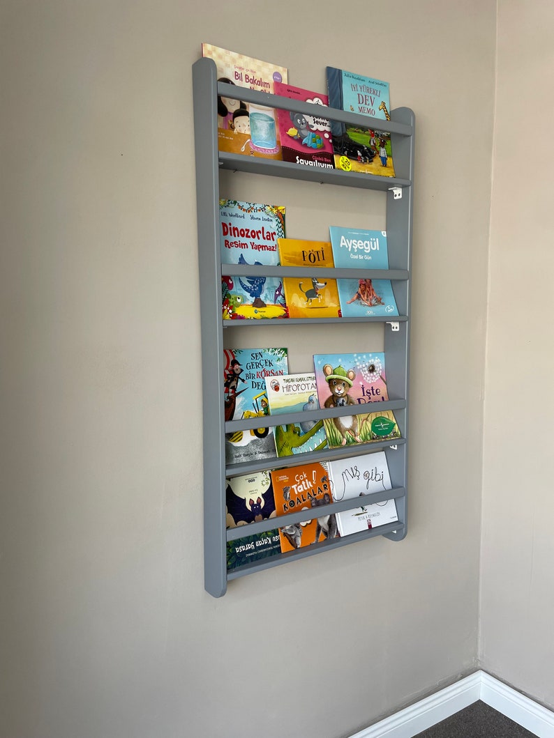 May include: A gray wooden bookshelf with multiple shelves, each holding a variety of colorful children's books. The books have titles in Turkish, including "Bil Bakalım", "Dinozorlar", "P&Ouml;TI", "Ayşeg&uuml;l", "SEN GER&Ccedil;EK BİR KORSAN DEG", "İşte", "&Ccedil;ok Tatlı KonalaLAR", and "us gibi".