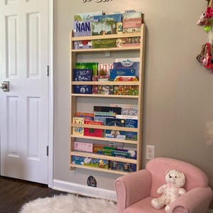 May include: A wooden bookshelf with multiple shelves filled with children's books. The bookshelf is mounted to the wall. A pink armchair with a teddy bear sitting on it is in the foreground. The wall behind the bookshelf has the words "once upon a time..." written on it.