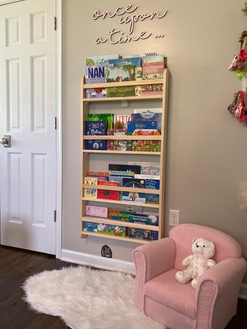 May include: A wooden bookshelf with multiple shelves filled with children's books. The bookshelf is mounted to the wall. A pink armchair with a teddy bear sitting on it is in the foreground. The wall behind the bookshelf has the words "once upon a time..." written on it.