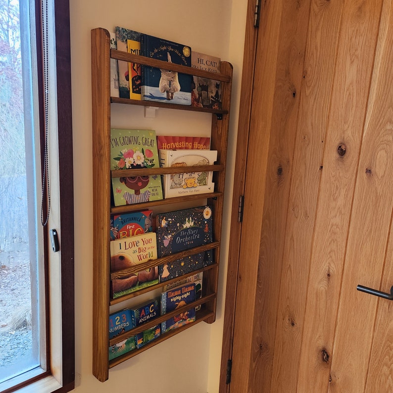 May include: A wooden bookshelf with five shelves, each holding a variety of children's books. The books have colorful covers with illustrations and titles such as "I'm Growing Great", "Harvesting Hope", "The Story Orchestra", and "ABC Animals".