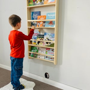 May include: A light wood wall-mounted bookshelf with multiple shelves filled with colorful children's books. A young boy is standing in front of the bookshelf, reaching for a book.