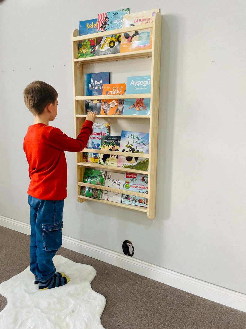 May include: A light wood wall-mounted bookshelf with multiple shelves filled with colorful children's books. A young boy is standing in front of the bookshelf, reaching for a book.