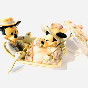 May include: A ceramic figurine of Mickey and Minnie Mouse sitting in a small boat. Mickey wears a straw hat and light suit, while Minnie wears a straw hat with pink flowers and a dress. Minnie holds a parasol decorated with pink floral accents.