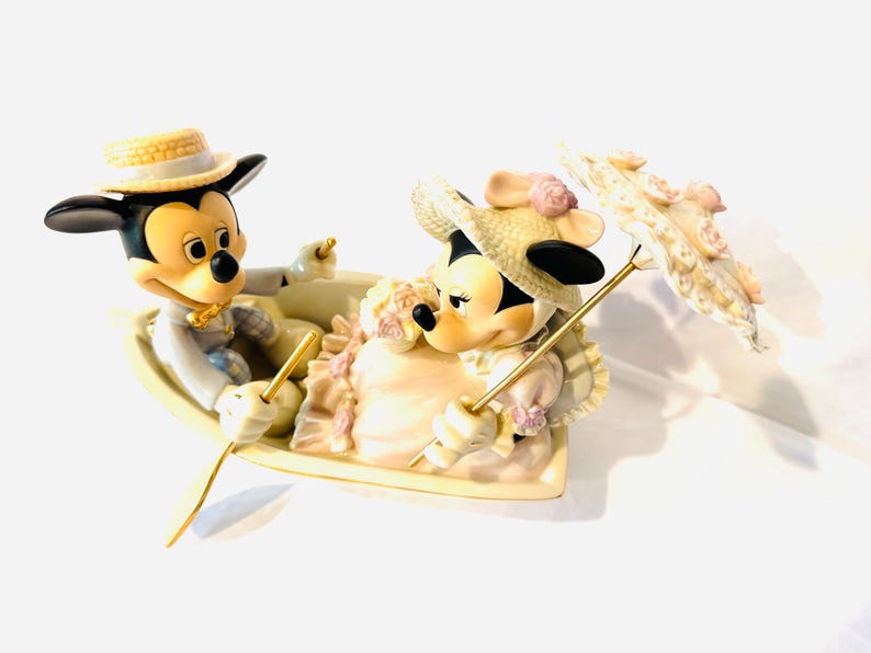 May include: A ceramic figurine of Mickey and Minnie Mouse sitting in a small boat. Mickey wears a straw hat and light suit, while Minnie wears a straw hat with pink flowers and a dress. Minnie holds a parasol decorated with pink floral accents.