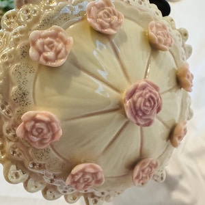 May include: A cream-colored ceramic lid featuring a glossy finish and a scalloped, lace-like edge. The top is decorated with several small, raised, light pink sculpted roses arranged in a circular pattern around a central rose.