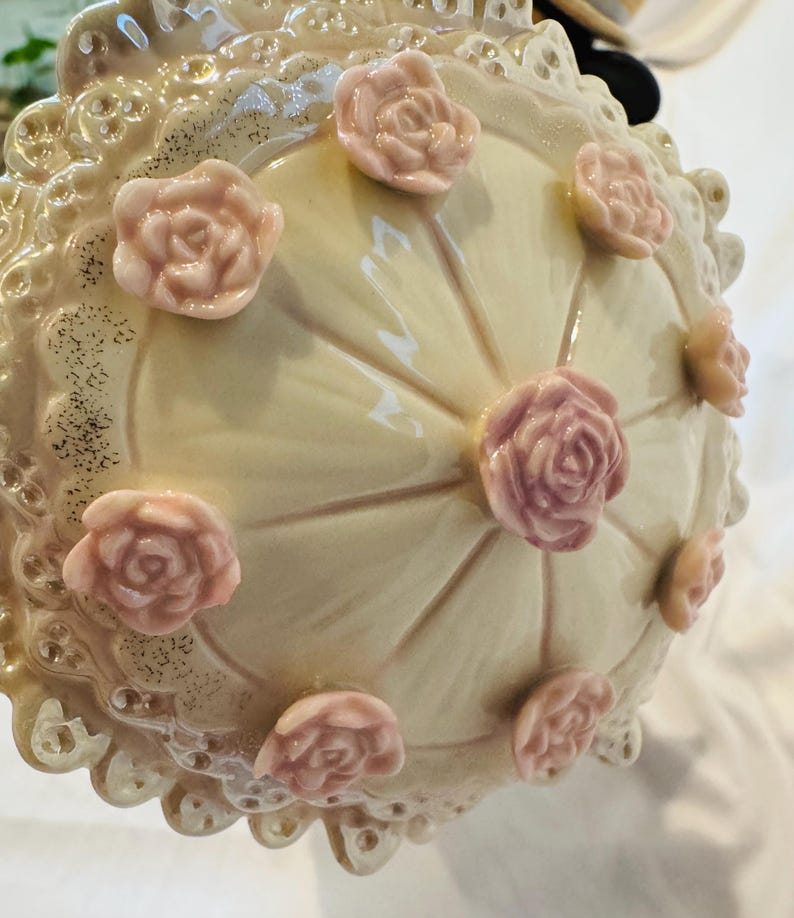 May include: A cream-colored ceramic lid featuring a glossy finish and a scalloped, lace-like edge. The top is decorated with several small, raised, light pink sculpted roses arranged in a circular pattern around a central rose.