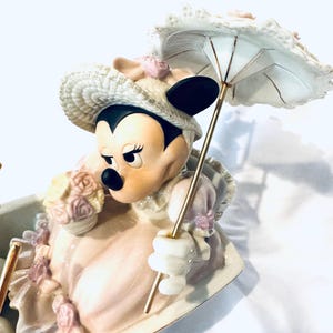 May include: A porcelain figurine of Minnie Mouse wearing a cream-colored bonnet and dress with pink floral accents. She holds a small bouquet of flowers and a white parasol with a gold-toned handle. The character is seated in a decorative vessel.