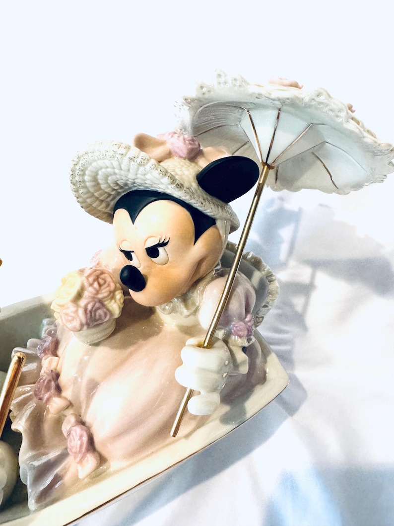 May include: A porcelain figurine of Minnie Mouse wearing a cream-colored bonnet and dress with pink floral accents. She holds a small bouquet of flowers and a white parasol with a gold-toned handle. The character is seated in a decorative vessel.