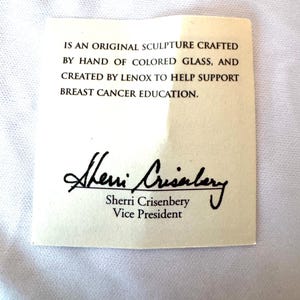 May include: A rectangular off-white paper card with black text that reads: IS AN ORIGINAL SCULPTURE CRAFTED BY HAND OF COLORED GLASS, AND CREATED BY LENOX TO HELP SUPPORT BREAST CANCER EDUCATION. It includes the signature of Sherri Crisenbery, Vice President.