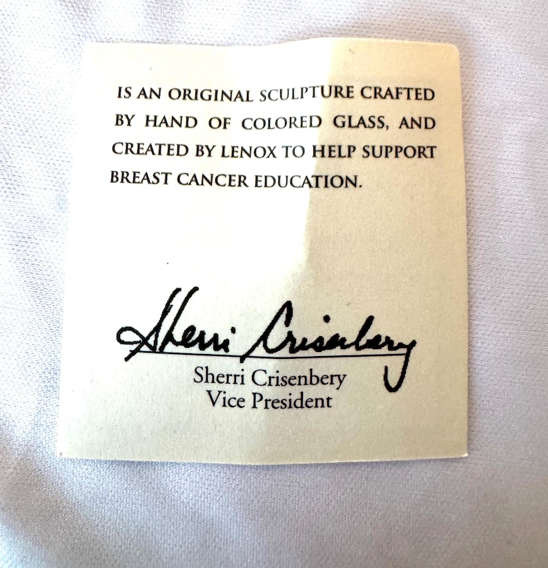 May include: A rectangular off-white paper card with black text that reads: IS AN ORIGINAL SCULPTURE CRAFTED BY HAND OF COLORED GLASS, AND CREATED BY LENOX TO HELP SUPPORT BREAST CANCER EDUCATION. It includes the signature of Sherri Crisenbery, Vice President.