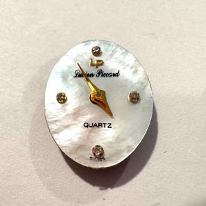 May include: Oval watch face with a mother-of-pearl background. The watch face features gold hands, the words "Lucien Piccard" and "QUARTZ" in black, and four small, sparkling accents. The word "SWISS" is at the bottom.