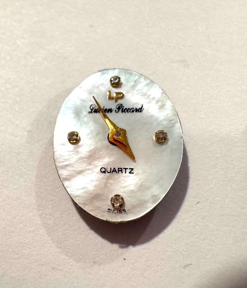 May include: Oval watch face with a mother-of-pearl background. The watch face features gold hands, the words "Lucien Piccard" and "QUARTZ" in black, and four small, sparkling accents. The word "SWISS" is at the bottom.
