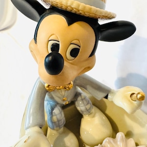 May include: A ceramic figurine of Mickey Mouse wearing a straw-colored boater hat, a light gray jacket, and plaid trousers. The character is seated in a small boat, holding a gold-colored oar, with a cluster of pale pink and white flowers in the front.