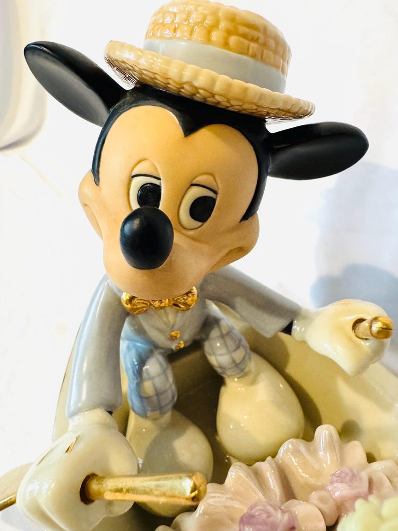 May include: A ceramic figurine of Mickey Mouse wearing a straw-colored boater hat, a light gray jacket, and plaid trousers. The character is seated in a small boat, holding a gold-colored oar, with a cluster of pale pink and white flowers in the front.