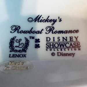 May include: A white ceramic base featuring dark blue text that reads Mickey's Rowboat Romance, Disney Showcase Collection, and Lenox. The logo includes a wreath surrounding the letter L, two clover icons, and a Made In China sticker at the bottom.
