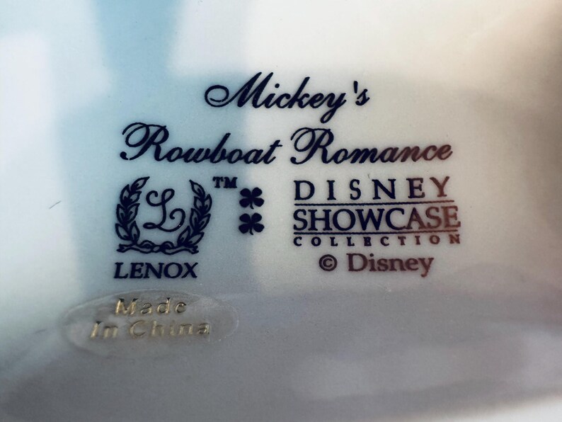 May include: A white ceramic base featuring dark blue text that reads Mickey's Rowboat Romance, Disney Showcase Collection, and Lenox. The logo includes a wreath surrounding the letter L, two clover icons, and a Made In China sticker at the bottom.