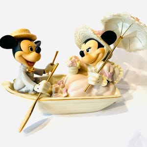 May include: A ceramic figurine of two cartoon mice in a small boat. The male mouse wears a straw hat and grey suit, rowing with a gold oar. The female mouse wears a straw hat, holds a bouquet of flowers, and carries a white lace parasol.