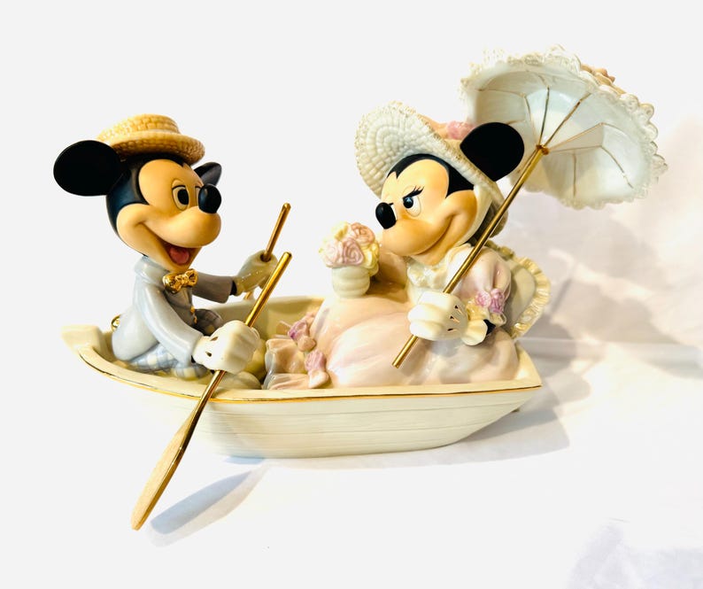 May include: A ceramic figurine of two cartoon mice in a small boat. The male mouse wears a straw hat and grey suit, rowing with a gold oar. The female mouse wears a straw hat, holds a bouquet of flowers, and carries a white lace parasol.