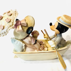 May include: A porcelain figurine of two cartoon mice in a small boat. One mouse wears a straw hat and holds a parasol with pink floral accents. The other mouse wears a straw boater hat and holds a gold-toned oar. The boat is cream-colored.