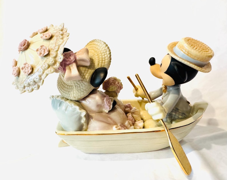 May include: A porcelain figurine of two cartoon mice in a small boat. One mouse wears a straw hat and holds a parasol with pink floral accents. The other mouse wears a straw boater hat and holds a gold-toned oar. The boat is cream-colored.